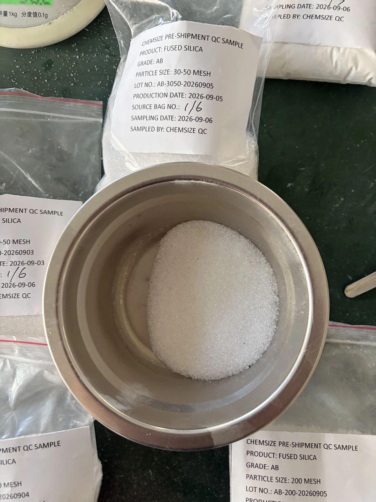 Close-up of white fused silica 30–50 mesh sand in a stainless steel weighing pan, with the labelled sample bag and part of a 500 g balance visible around it.