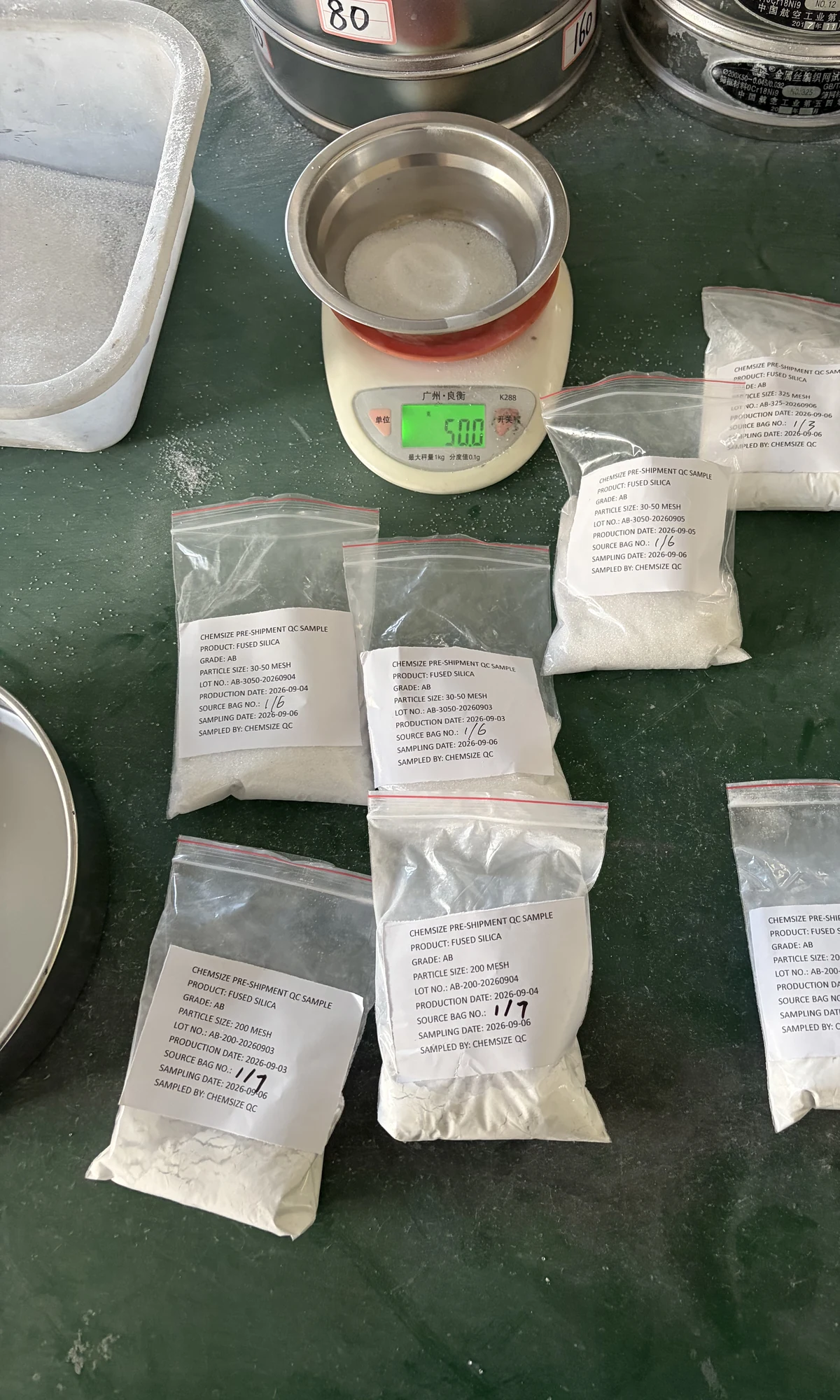 A 500 g laboratory balance and a grid of labelled sample bags of fused silica powder laid out on a bench, with sieve pans at the edge of the frame, during a pre-shipment quality check.