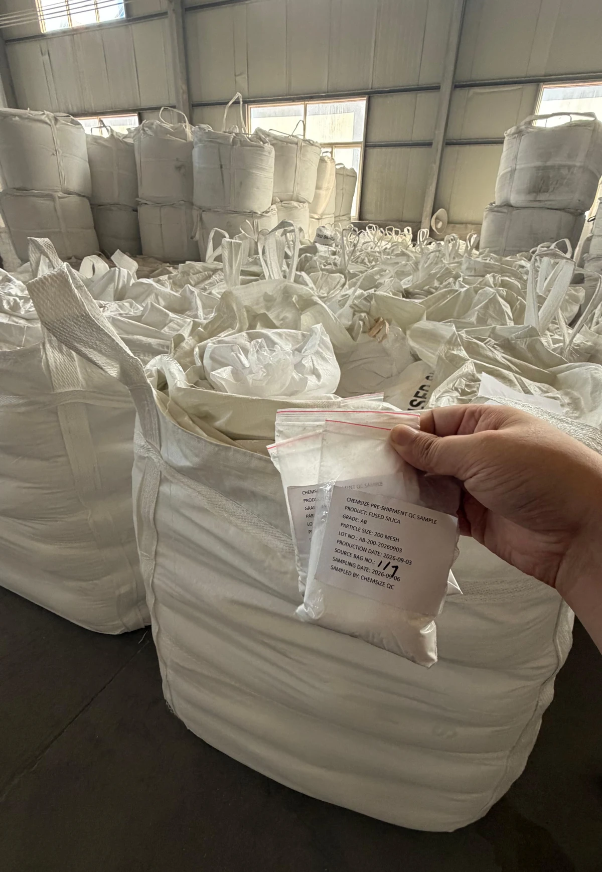 A hand holding a small labelled sample bag of fused silica powder over an open FIBC jumbo bag, with rows of filled jumbo bags stacked in the warehouse behind.
